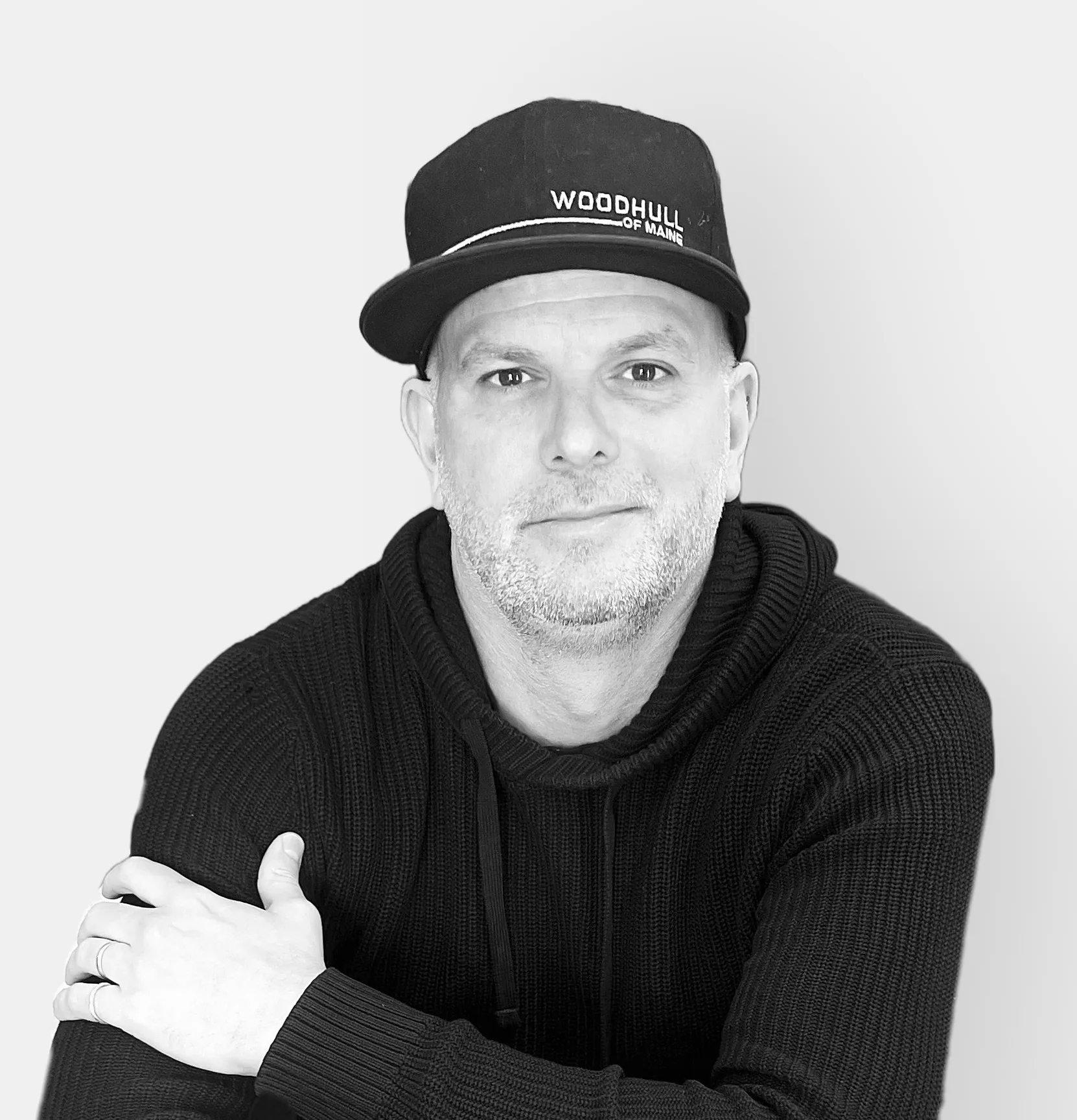 A man in a black hoodie and cap sits with one arm resting on his knee, smiling slightly against a plain, light background.
