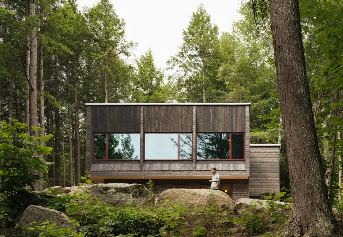 Pieri Pines modern wood cabin with large reflective windows nestled among tall pine trees and boulders in a forest clearing