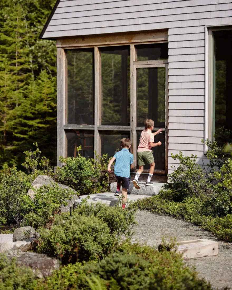 Two boys running towards, Pine Cove, a modern cabin with gray siding and large windows, surrounded by green foliage and a stone path