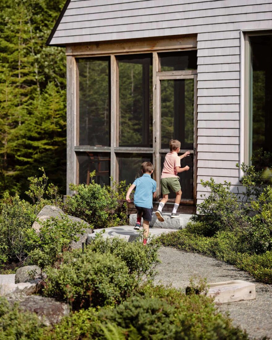 Two boys running towards, Pine Cove, a modern cabin with gray siding and large windows, surrounded by green foliage and a stone path