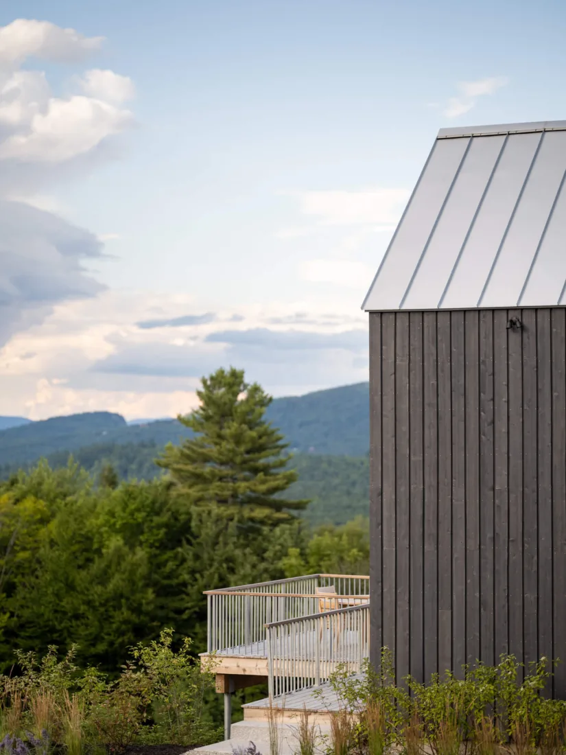 A modern dark wooden building with a metal roof, designed by an architect, sits by a wooden deck surrounded by trees and mountains under a partly cloudy sky.