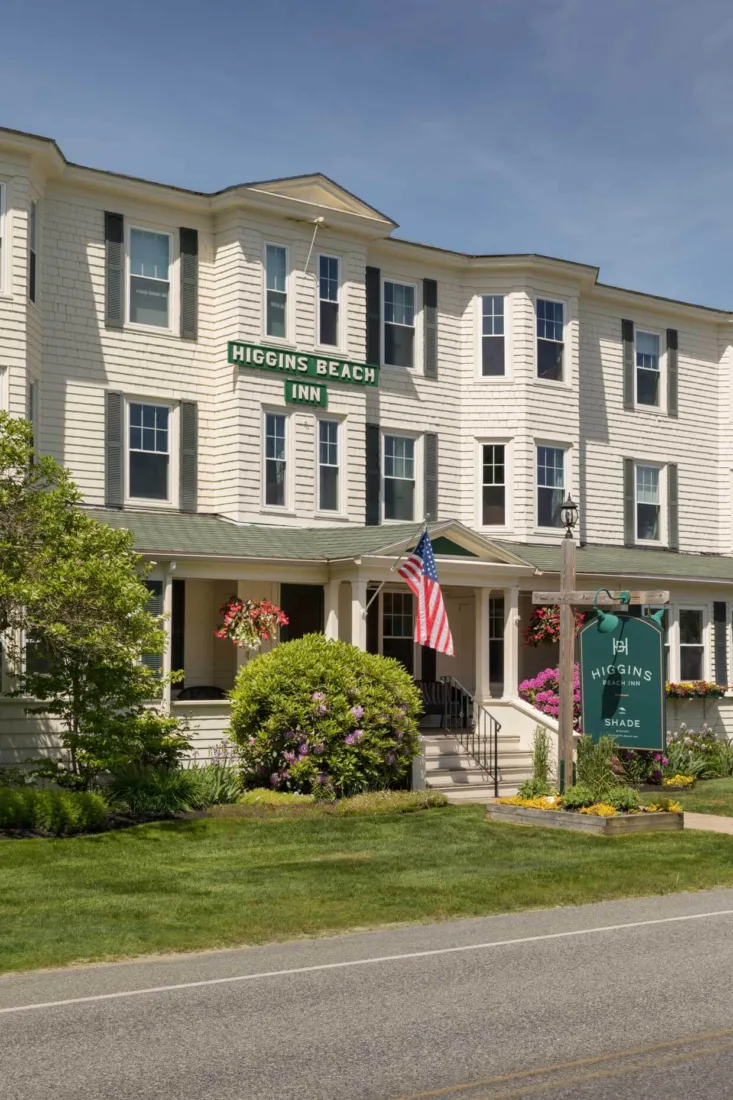 A three-story white clapboard inn designed by local architect s, with green “Higgins Beach Inn” signs, a flower-lined porch, and an American flag greeting guests on a sunny day.