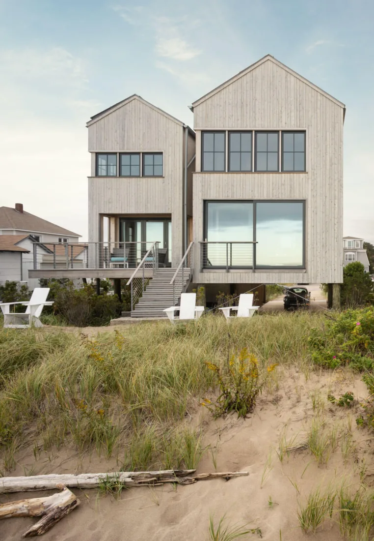 Two modern, light wood beach houses on stilts by a Maine architect face sandy dunes; white outdoor chairs are arranged on the sand among tall grasses.
