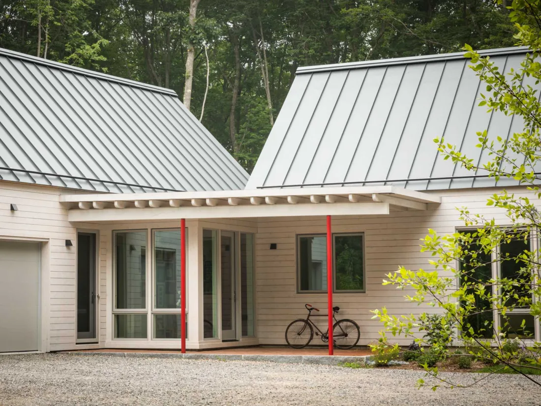 Modern house by Maine homebuilders with light wood siding, metal roof, red entry posts, large windows, a bicycle outside, and nestled among trees.