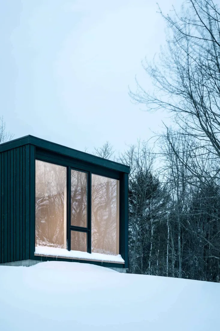 A modern black cabin designed by a Maine architect sits on a snowy hill, surrounded by bare winter trees under a pale sky.