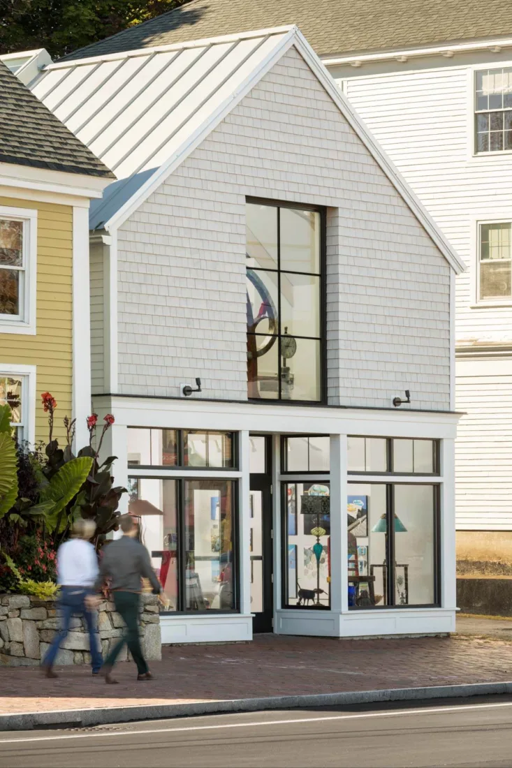 A modern glass-fronted art gallery by a Maine architect, with white shingles and large windows, stands between two traditional buildings as two people walk past on the sidewalk.