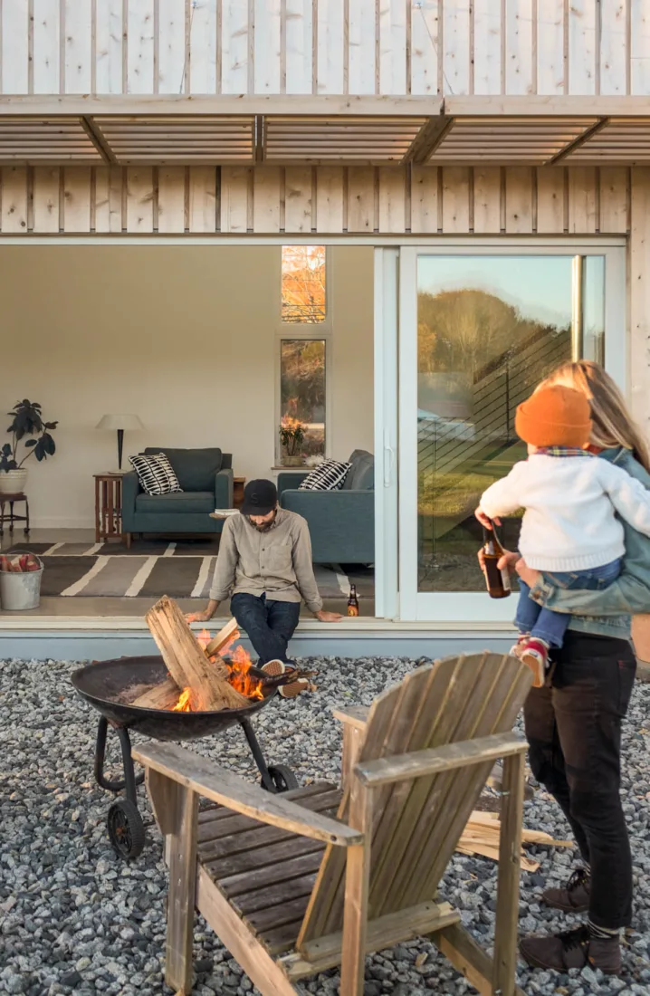 A man sits by an outdoor fire pit near a modern house by Maine homebuilders as a woman holds a child and drink, with wooden chairs and a cozy living room visible inside.