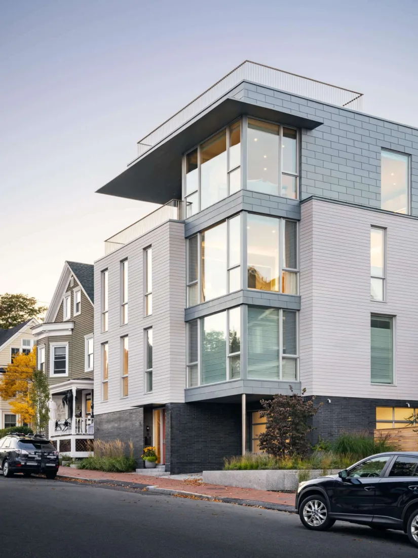 Modern multi-story building with large glass windows and gray siding, designed by a Maine architect, sits on a city street next to older houses with cars parked along the sidewalk.