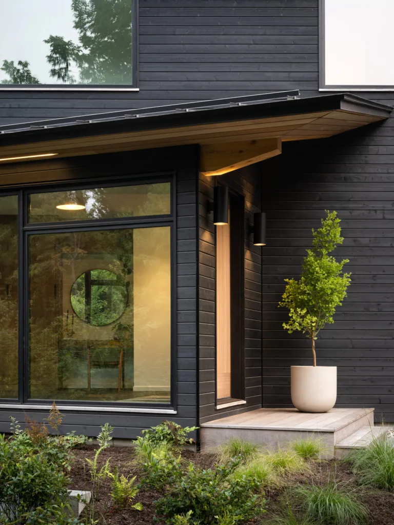 Striking front entryway of a custom luxury home in Maine, featuring sleek black siding, a circular interior window, and intentional landscaping