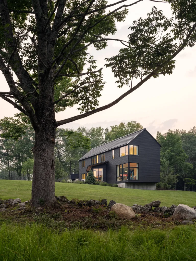 Modern luxury design-build home in New Gloucester, Maine, with large black-framed windows glowing at sunset and surrounded by natural New England landscapes