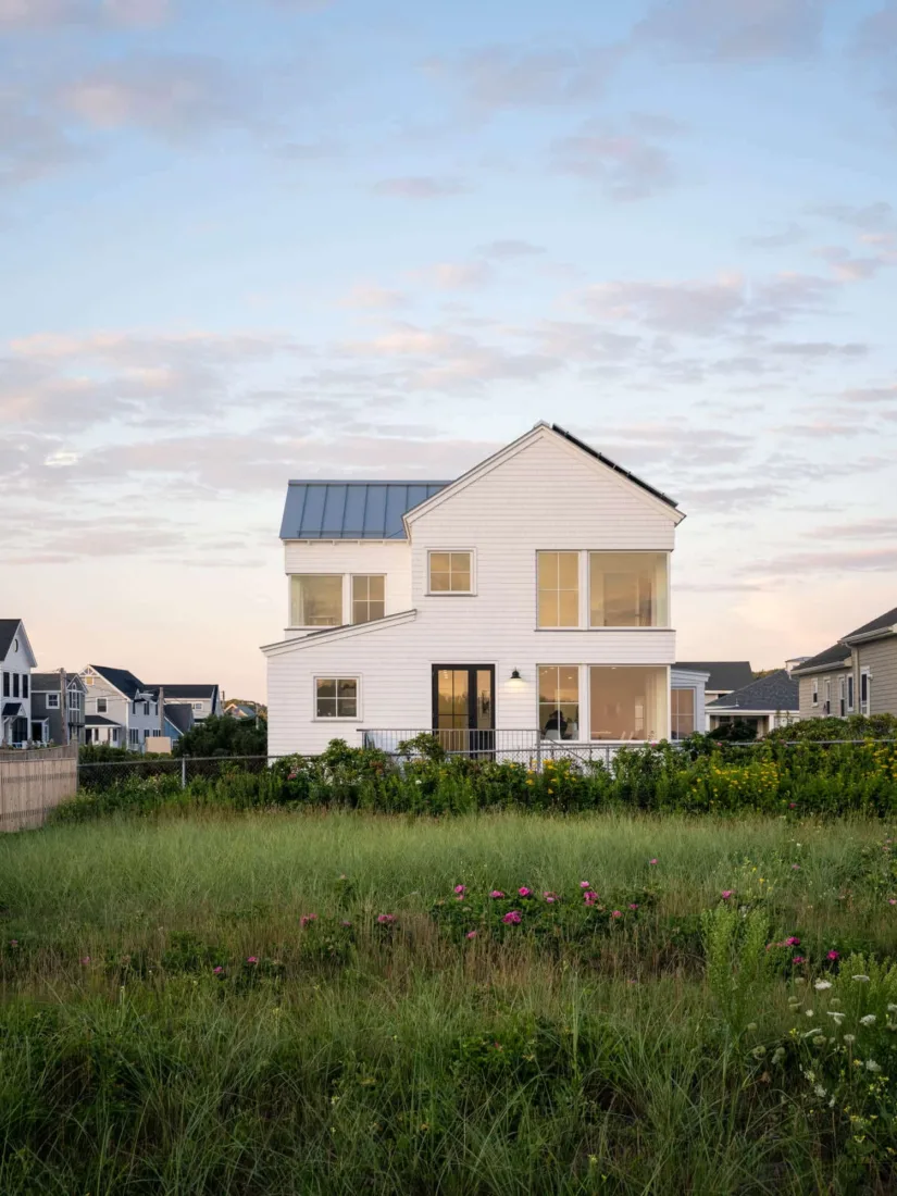 A modern white two-story house with large windows and a slanted roof, crafted by Maine homebuilders, sits amid green grass and wildflowers under a partly cloudy sky.