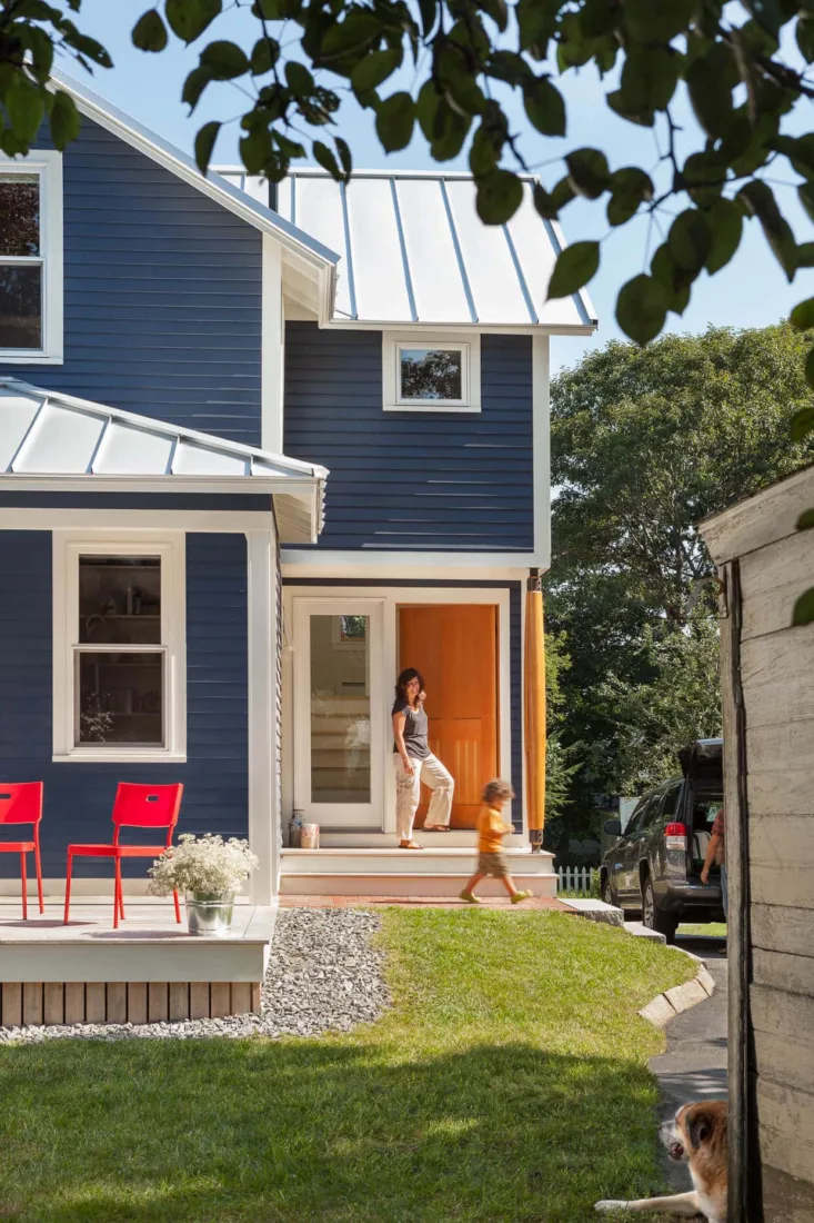A woman stands at the door of a blue house by Maine homebuilders as a child runs down the steps. Two red chairs sit on the porch, and a dog is partly visible in the yard.