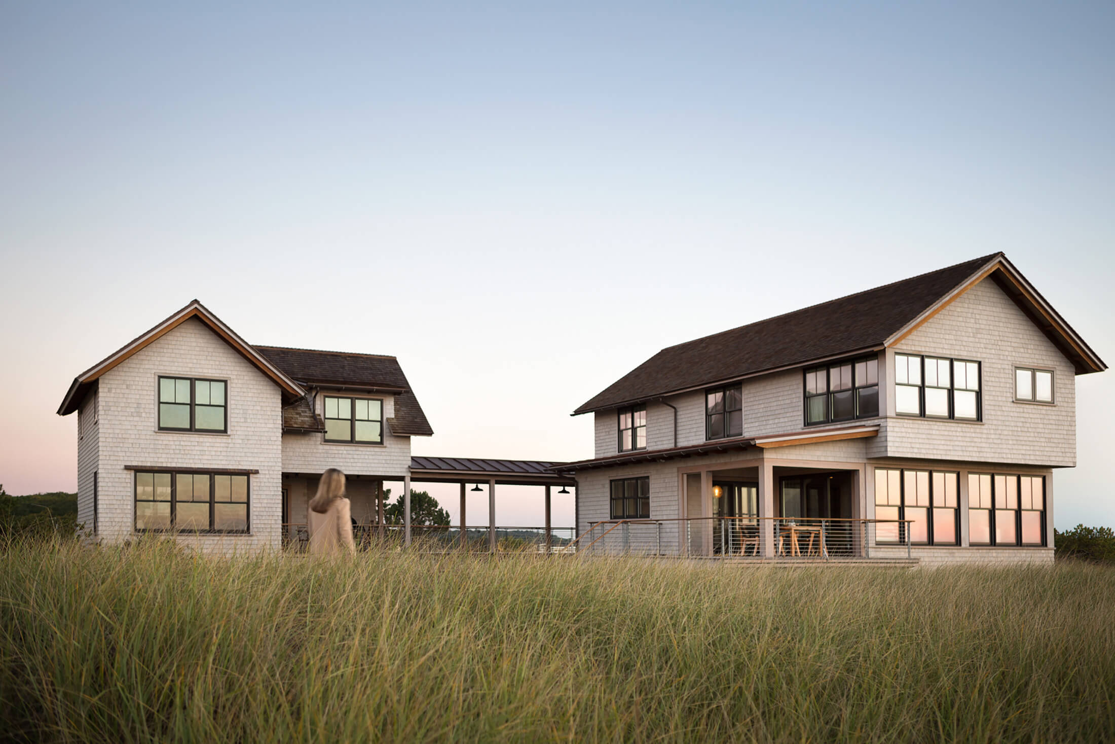 Exterior view of Cast Away house Project. Long grass with a woman walking towards the house with many windows and an exterior covered walk way spanning between the house