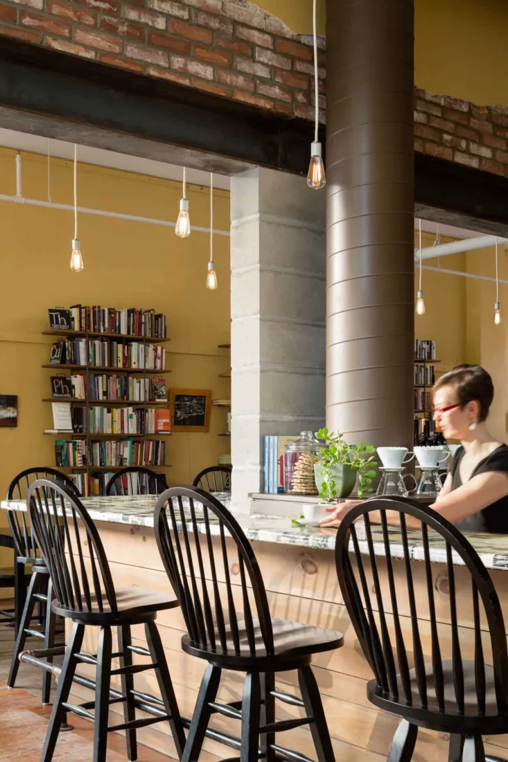 A cozy café with wooden chairs at a counter, bookshelves in the background, hanging lights, and a Maine architect thoughtfully preparing coffee.