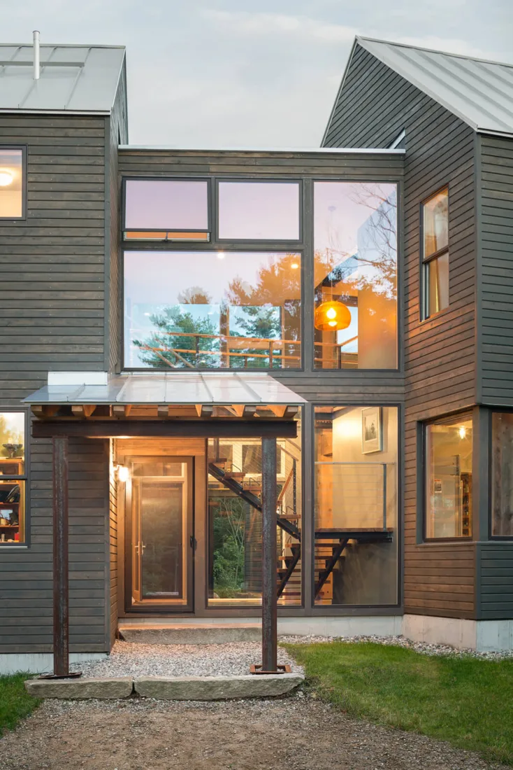 Modern two-story house by Maine homebuilders with large glass windows, gray wood siding, visible staircase, warm interior lighting, and a gravel entryway with a wooden pergola.