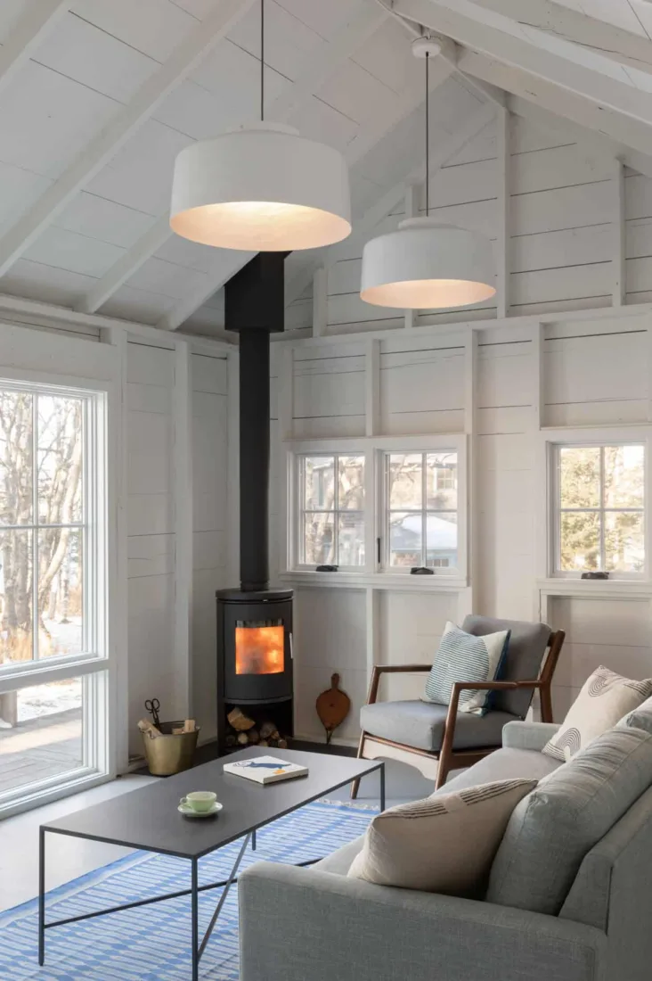 A cozy, modern living room in true Maine homebuilders style, featuring a wood-burning stove, gray sofa, blue rug, and pendant lights under bright white walls.