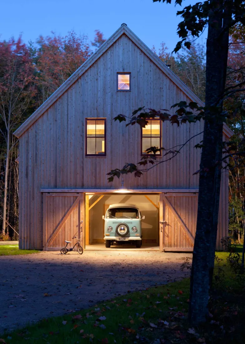 A vintage blue van is parked in a barn-style garage designed by a Maine architect; warm lights glow from the windows as dusk falls, with a tricycle nearby.
