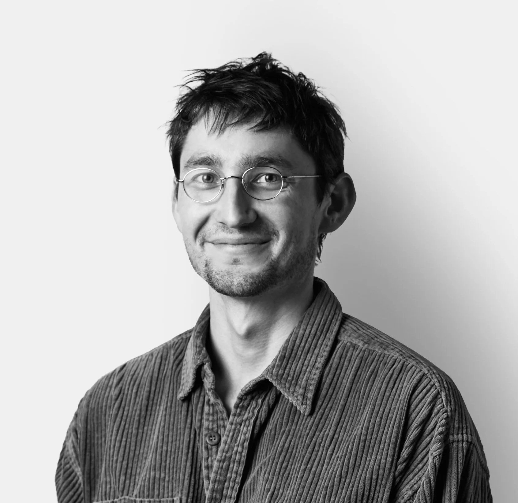 A young man with glasses and short hair, wearing a corduroy shirt, smiles while standing against a plain white background.