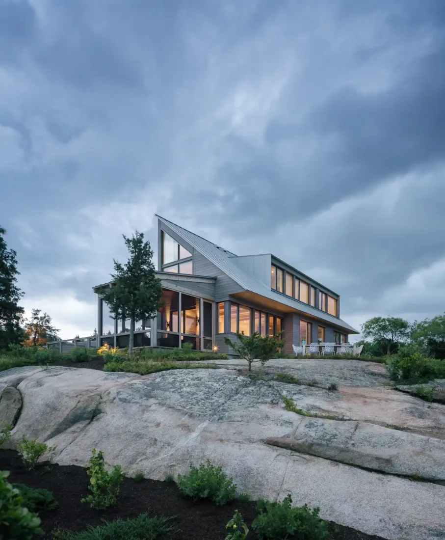 Modern two-story house with large windows and angled roof, set on rocky terrain with greenery—showcasing design-build expertise under a cloudy evening sky.