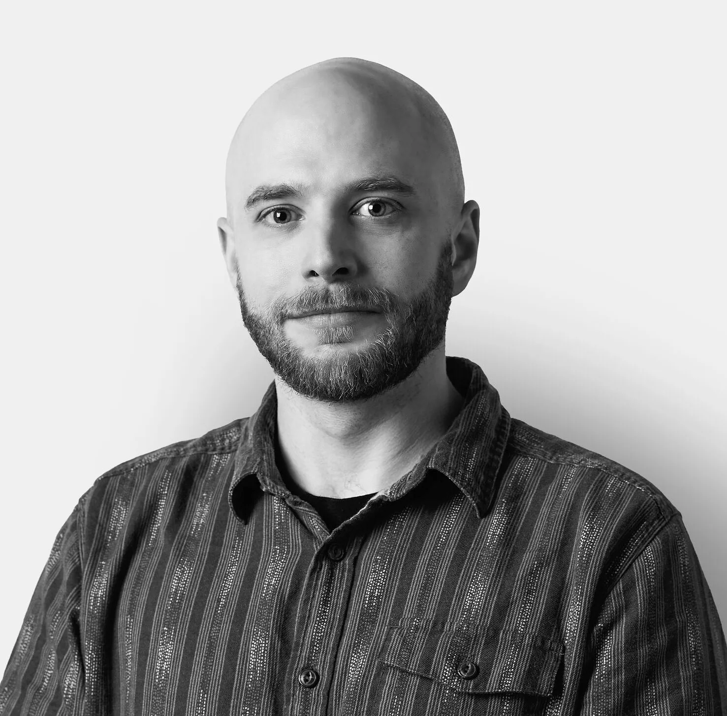 A bald man with a beard wearing a striped button-up shirt stands against a plain white background, looking at the camera with a neutral expression.