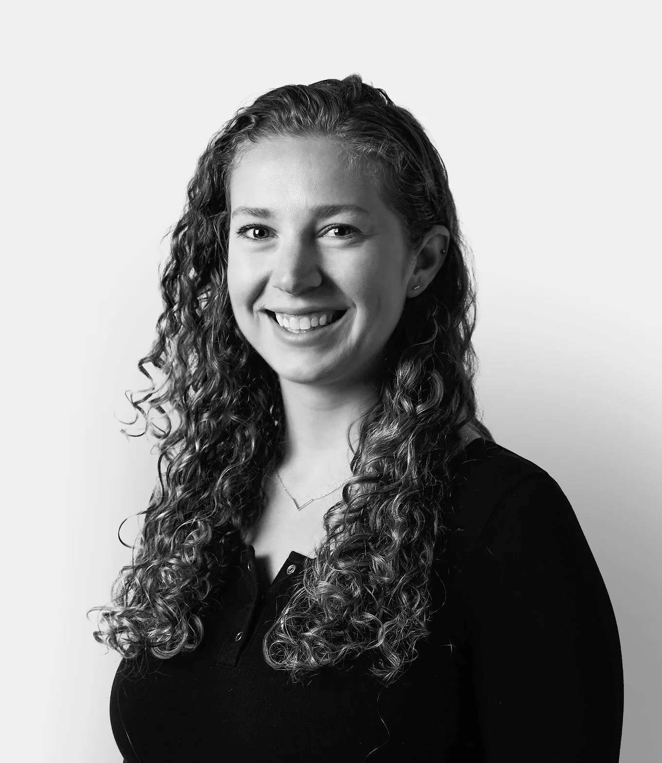 A woman with long curly hair, wearing a black long-sleeve top, smiles at the camera against a plain light background.