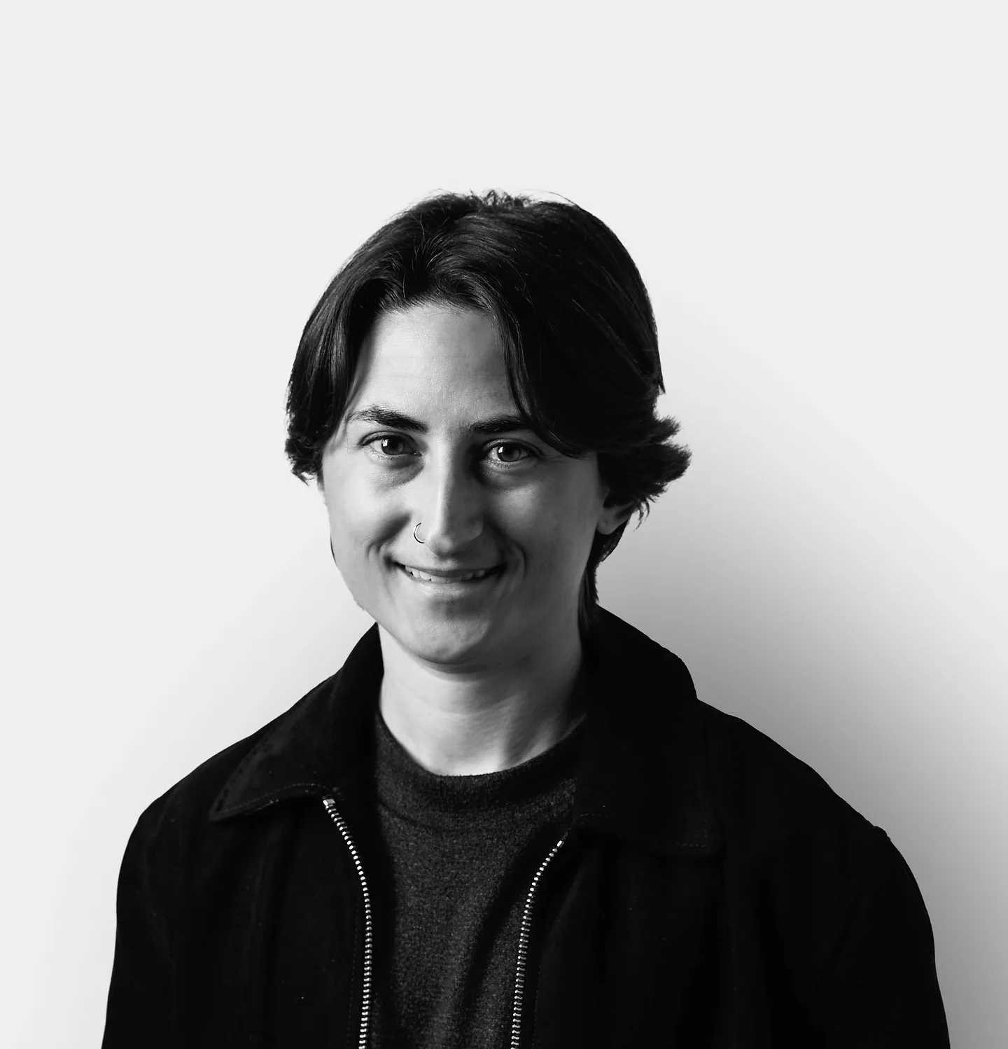 A person with short, dark hair smiles at the camera, wearing a dark jacket over a shirt, standing against a plain white background.