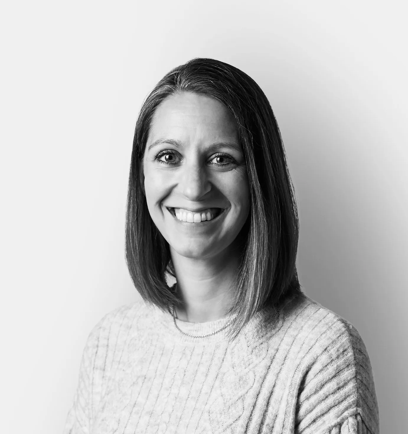 A woman with straight shoulder-length hair wearing a light-colored, cable-knit sweater smiles at the camera against a plain background.
