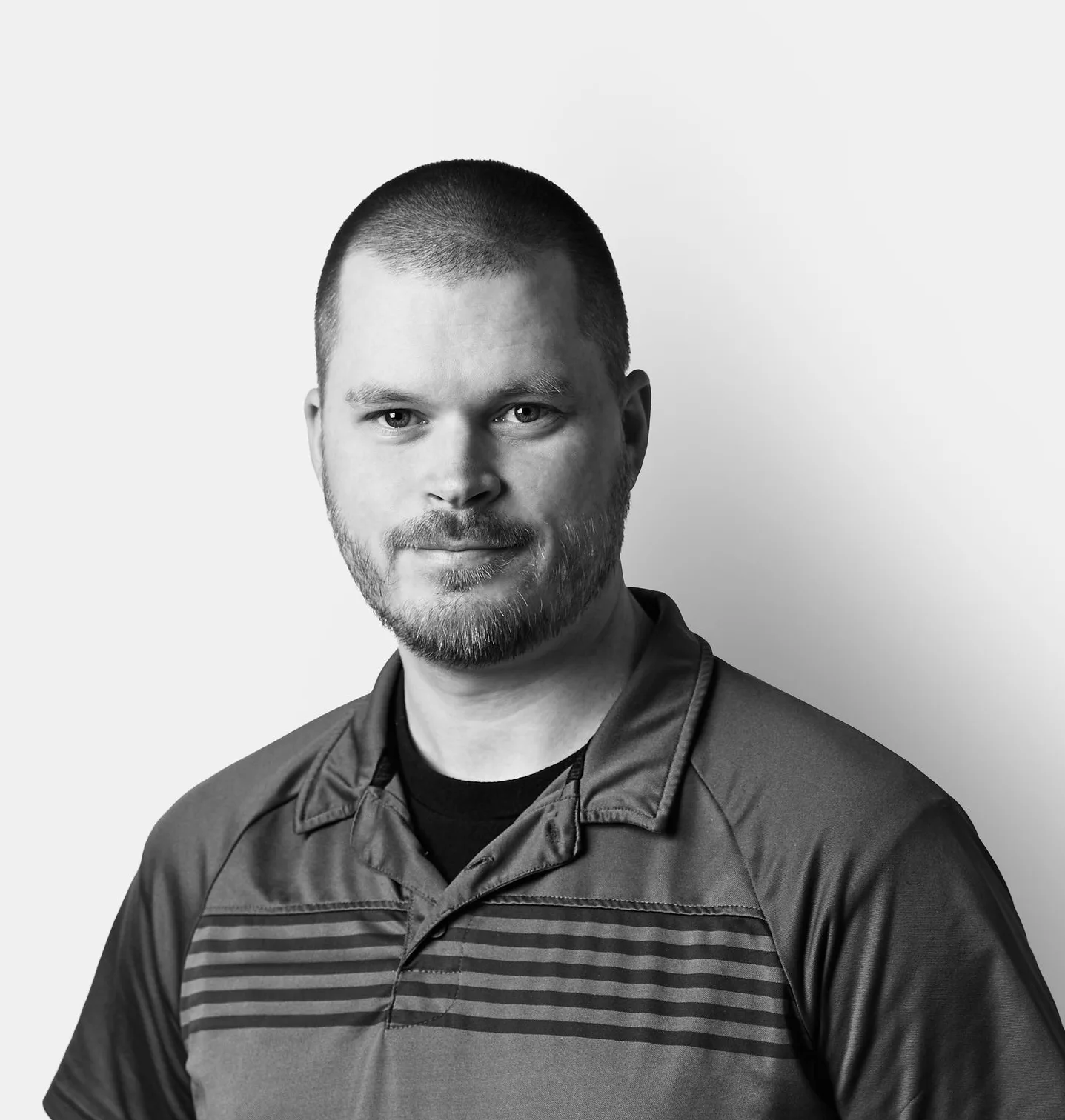 A man with short hair and a beard, wearing a short-sleeved polo shirt, stands against a plain, light background and looks at the camera.