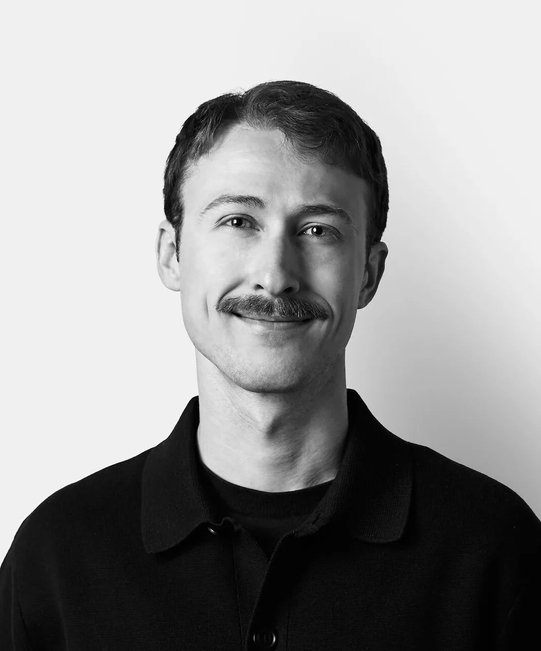 A man with short hair and a mustache smiles slightly, wearing a dark buttoned shirt, standing against a plain white background.