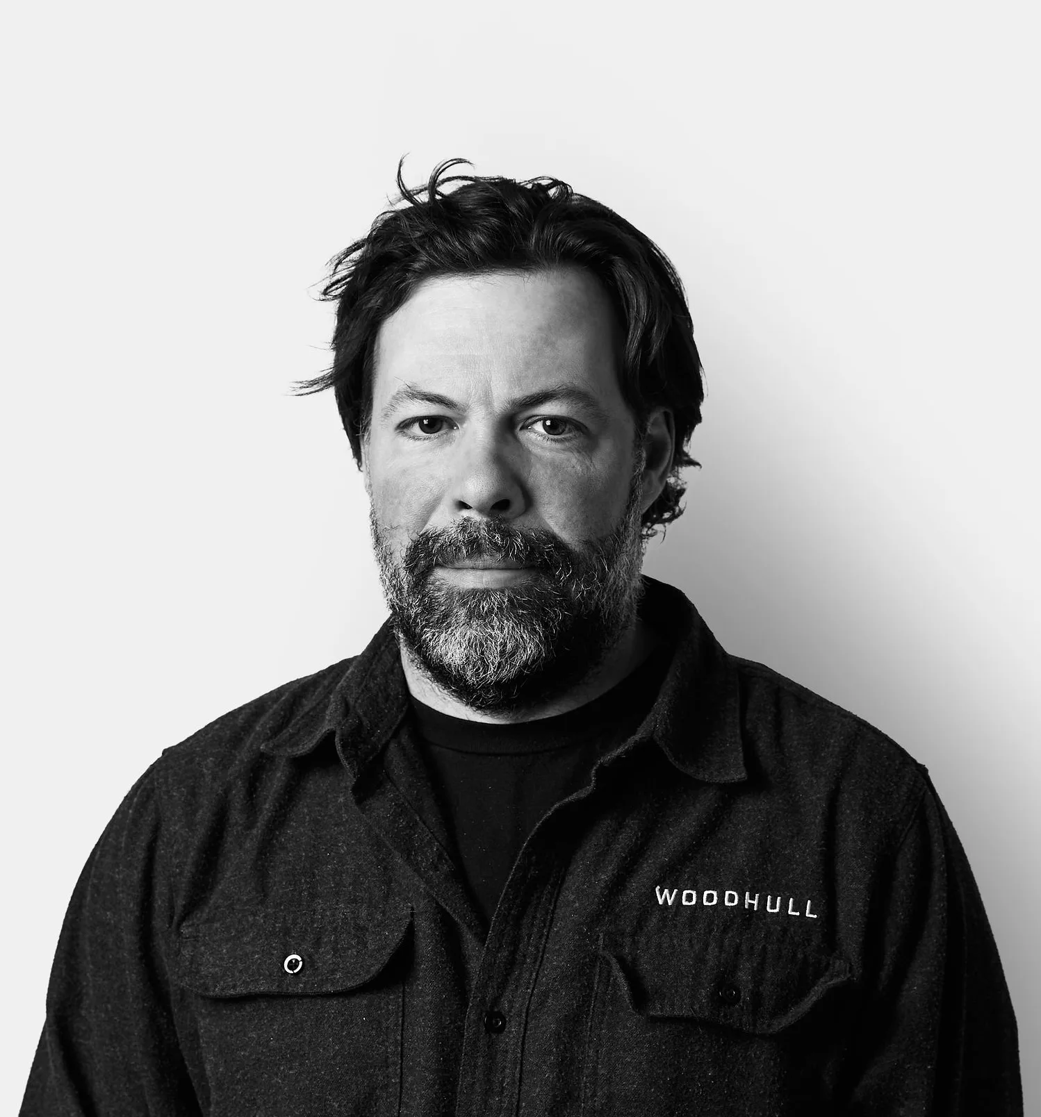 A man with a beard and mustache wearing a dark shirt that says WOODHULL stands against a plain white background, looking directly at the camera.