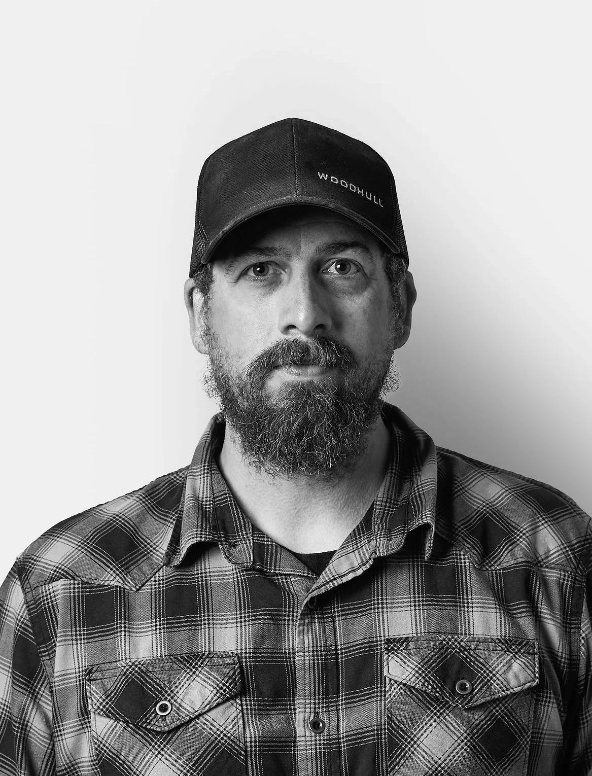 A man with a beard wearing a plaid shirt and a dark baseball cap stands against a plain white background, looking directly at the camera.