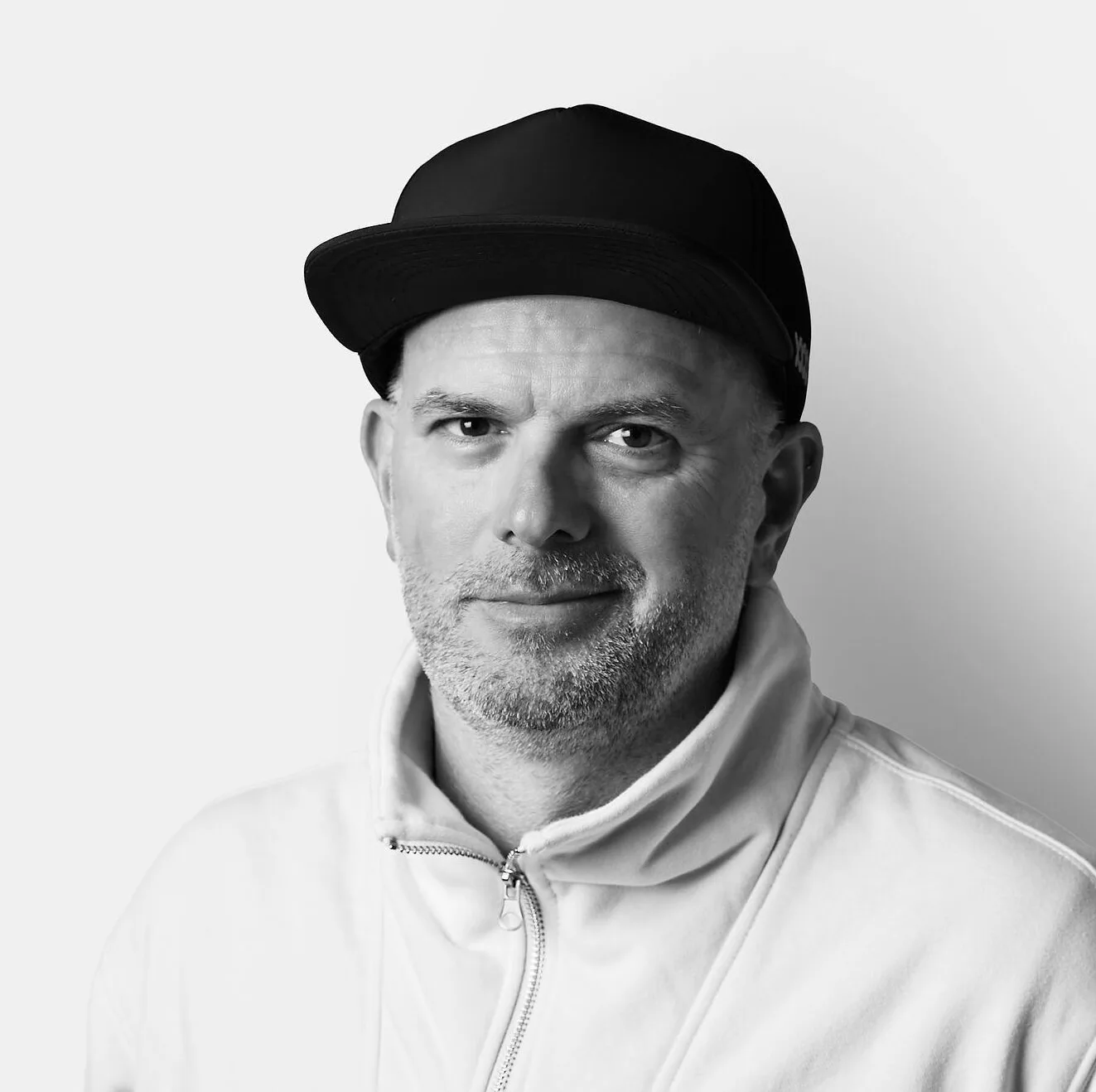 A man with a beard wearing a black cap and a white zip-up sweater stands against a plain white background, looking at the camera with a neutral expression.