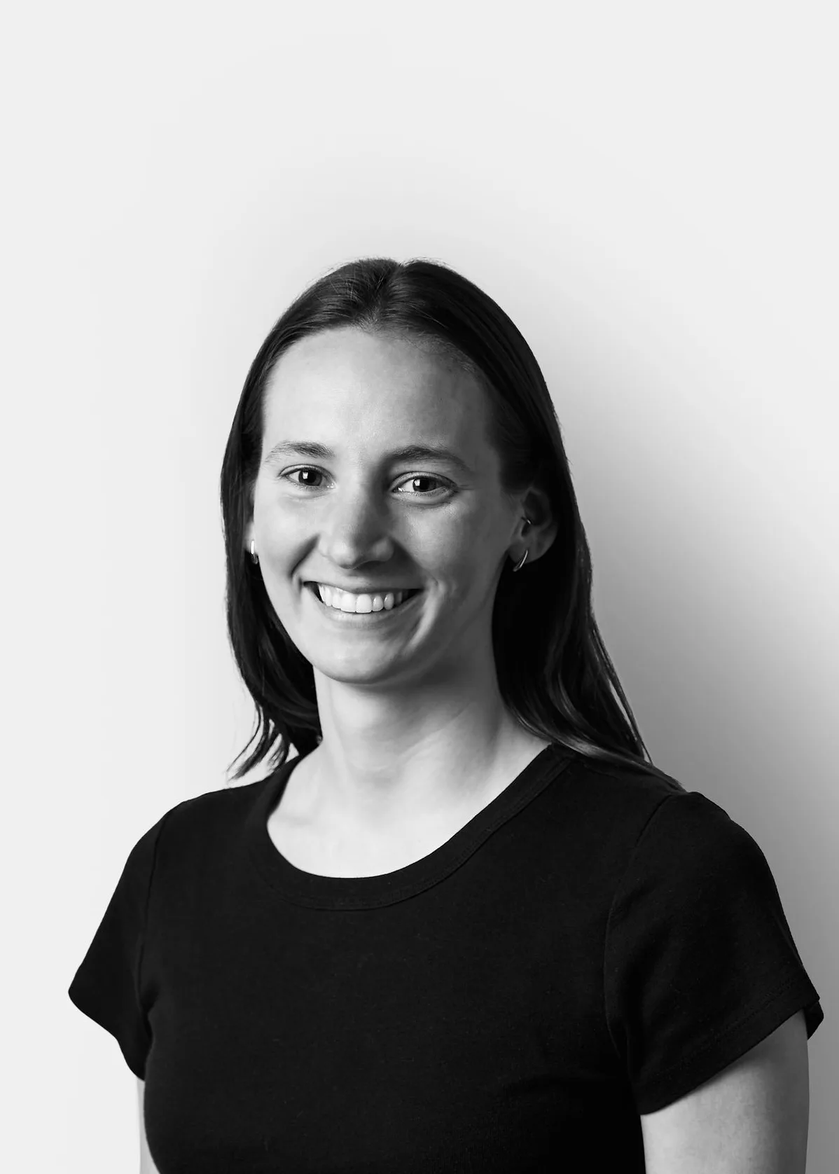 A woman with long dark hair, wearing a black short-sleeve top, smiles while standing against a plain white background. The photo is in black and white.