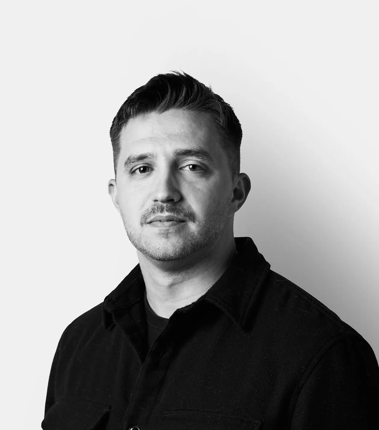 A man with short hair and a mustache, wearing a dark button-up shirt, stands against a plain white background, looking at the camera with a neutral expression.