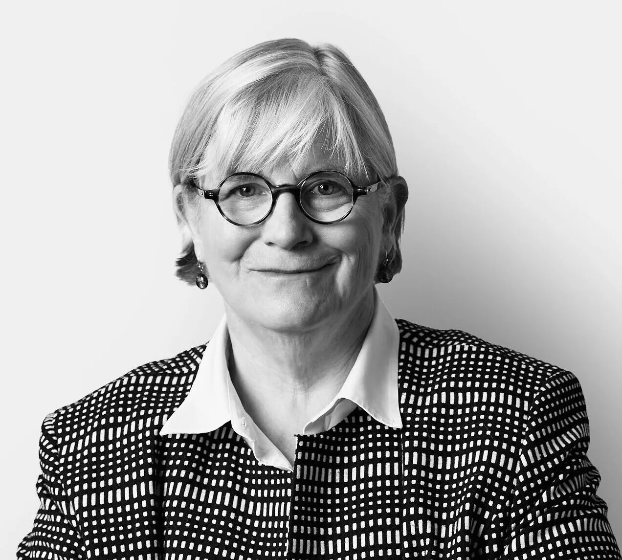 A person with short, light hair and glasses wearing a patterned jacket and a collared shirt, smiling and posed against a plain white background.
