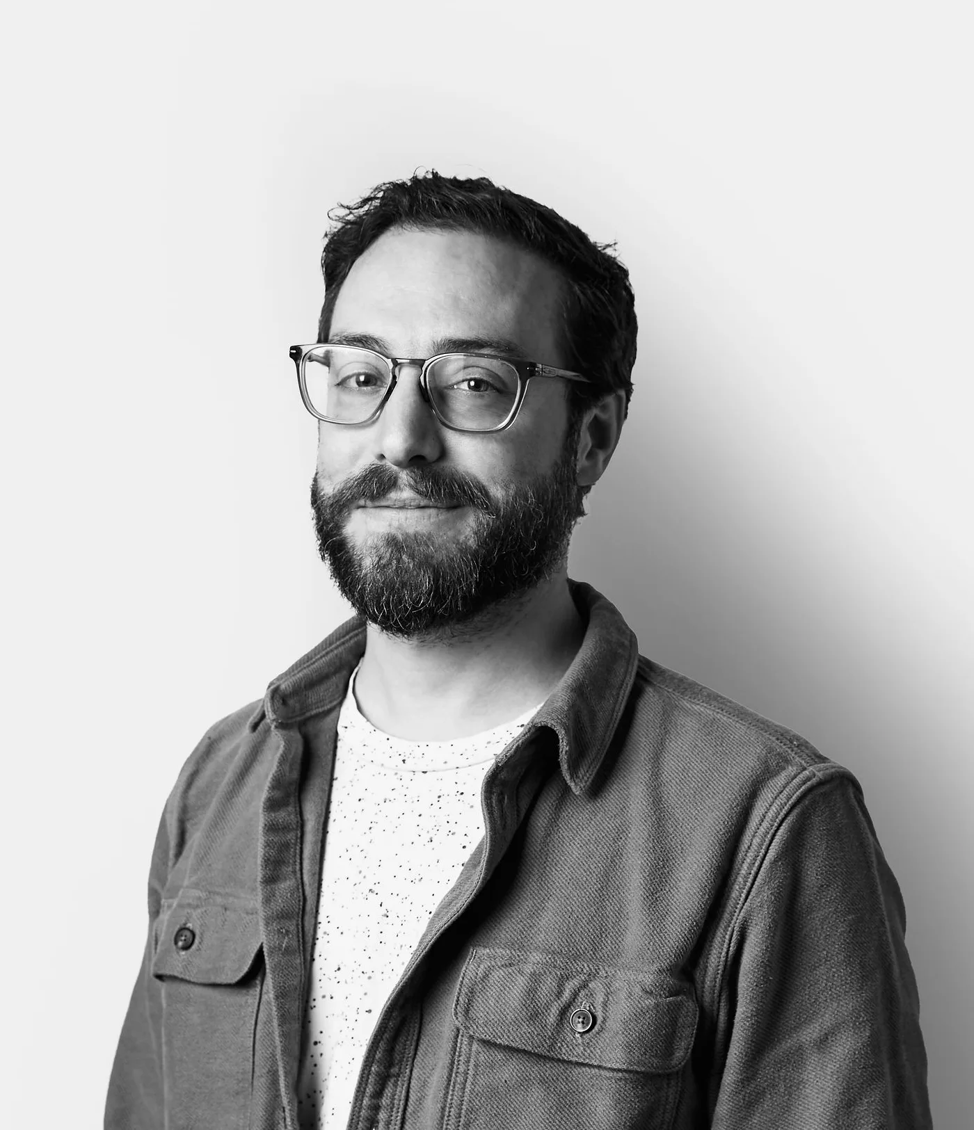 A man with glasses and a beard, wearing a light shirt and a jacket, stands against a plain white background, looking at the camera with a slight smile.