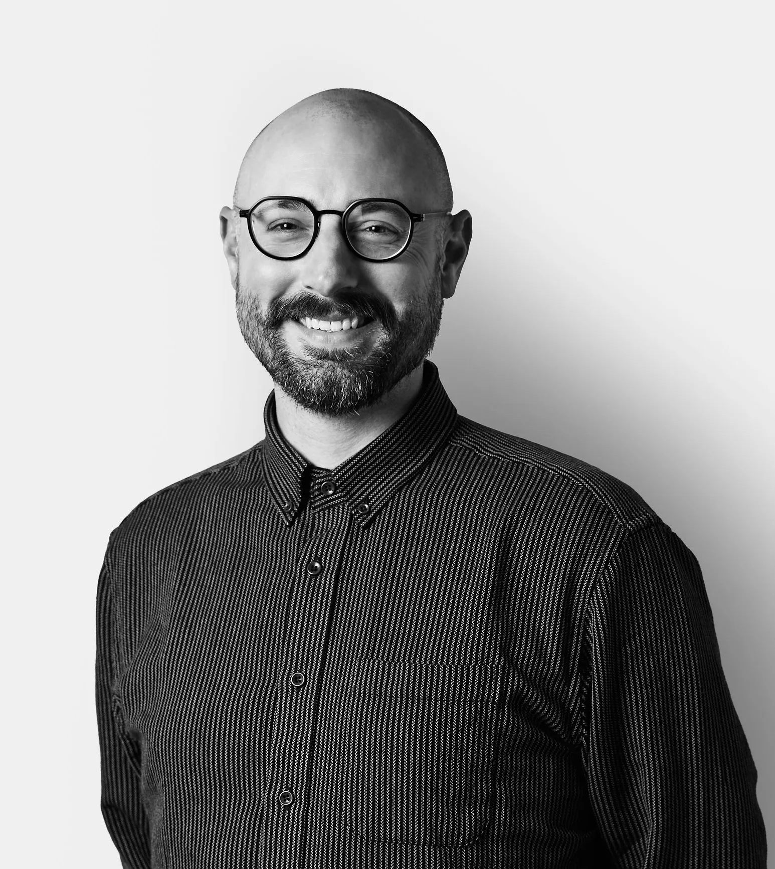A smiling man with a beard, shaved head, and glasses wears a dark, striped button-up shirt, standing against a plain white background.