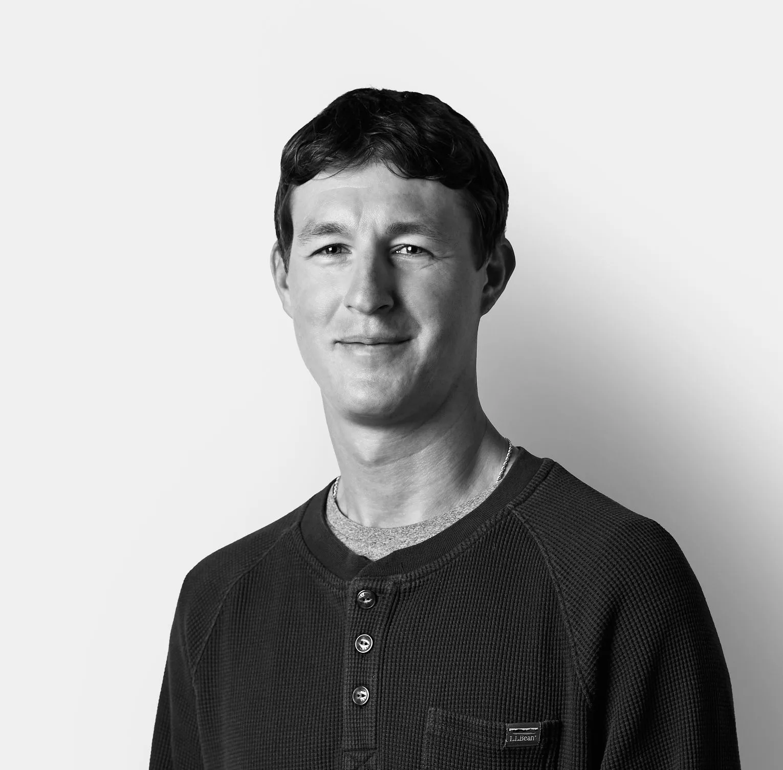 A man with short dark hair wearing a long-sleeve, dark henley shirt stands against a plain, light background, smiling slightly.