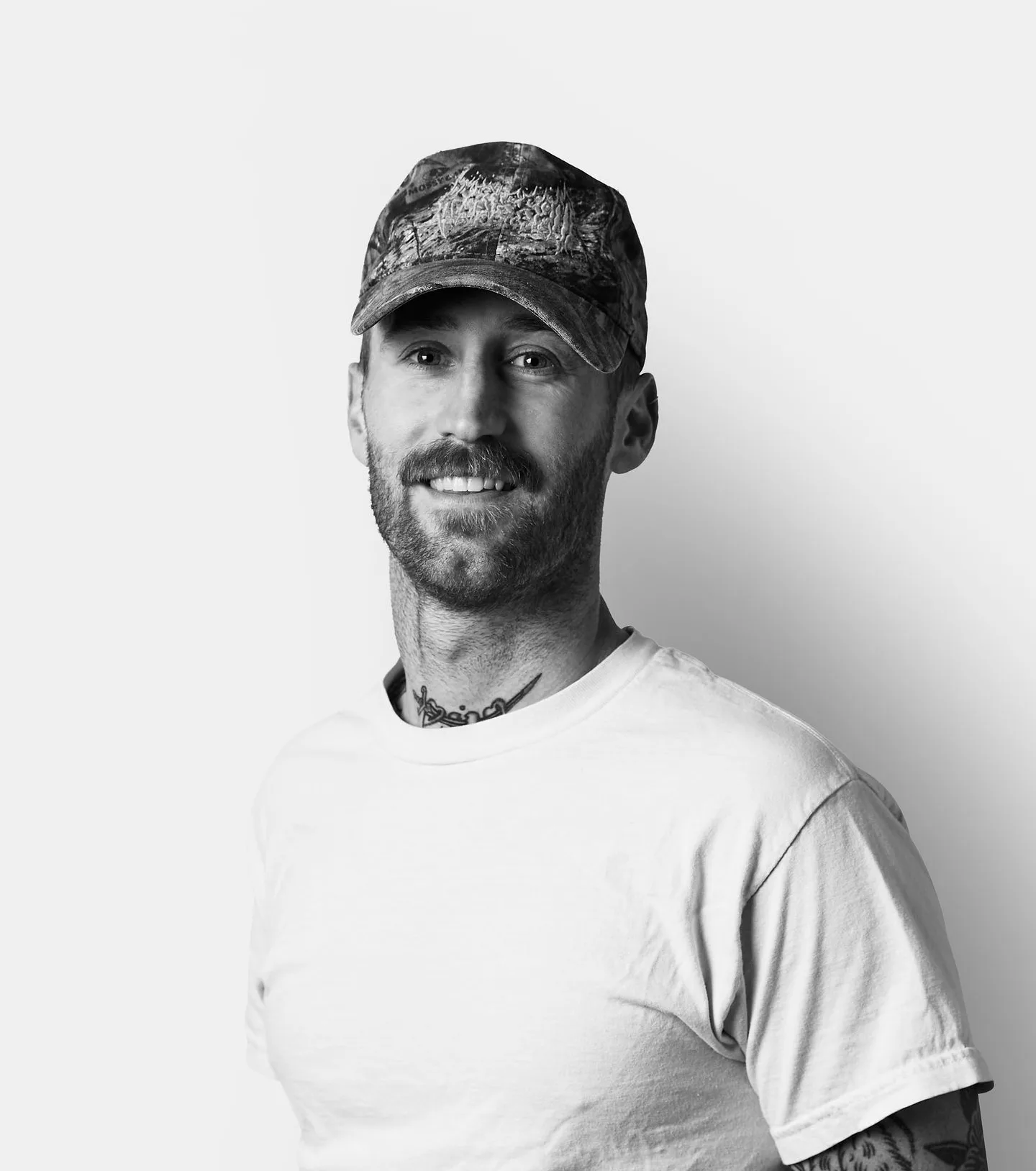 Man with tattoos wearing a patterned cap and plain t-shirt, smiling at the camera against a plain, light background.