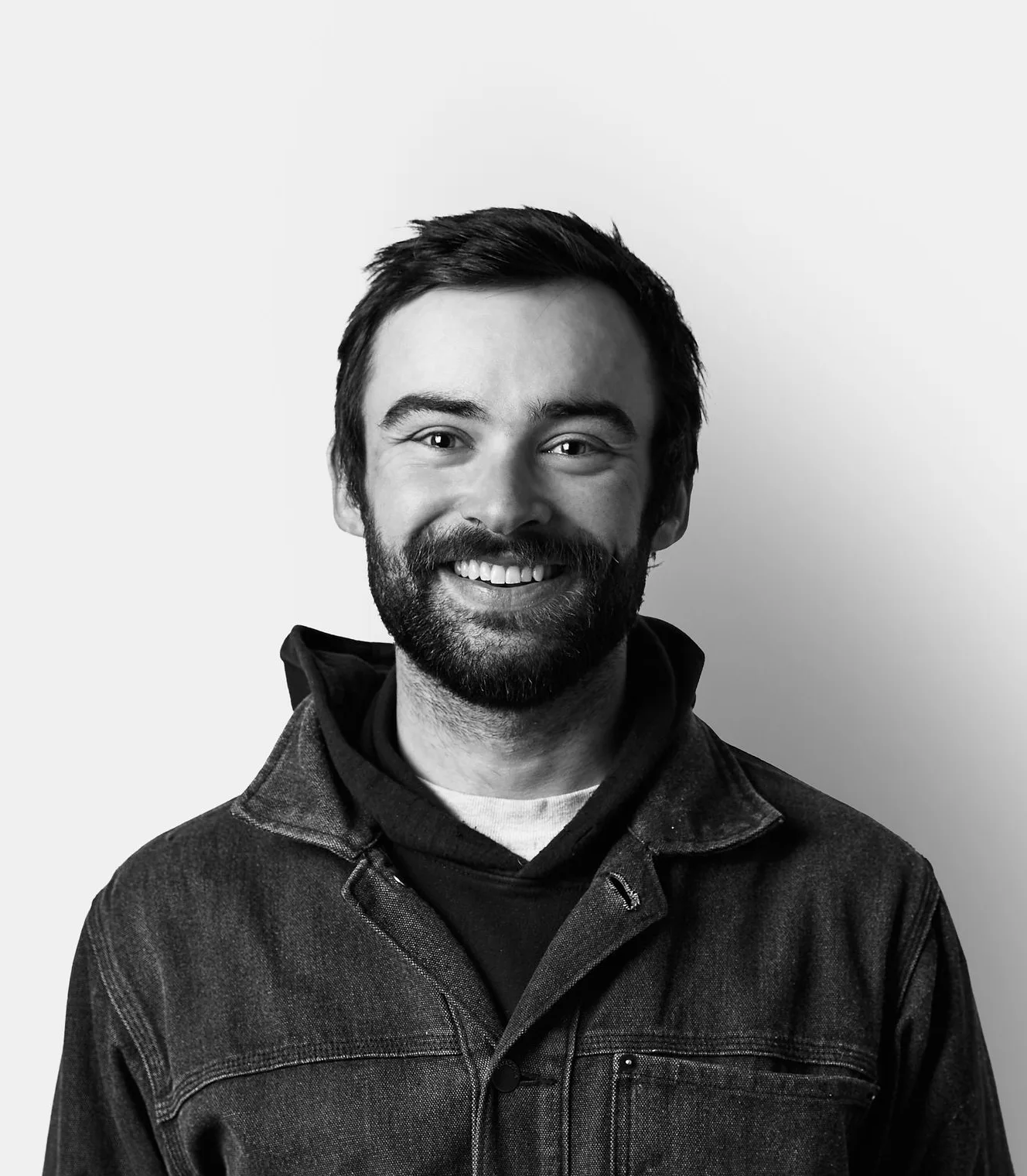 A man with a beard and dark hair smiles at the camera. He is wearing a denim jacket over a hoodie, standing against a plain white background.