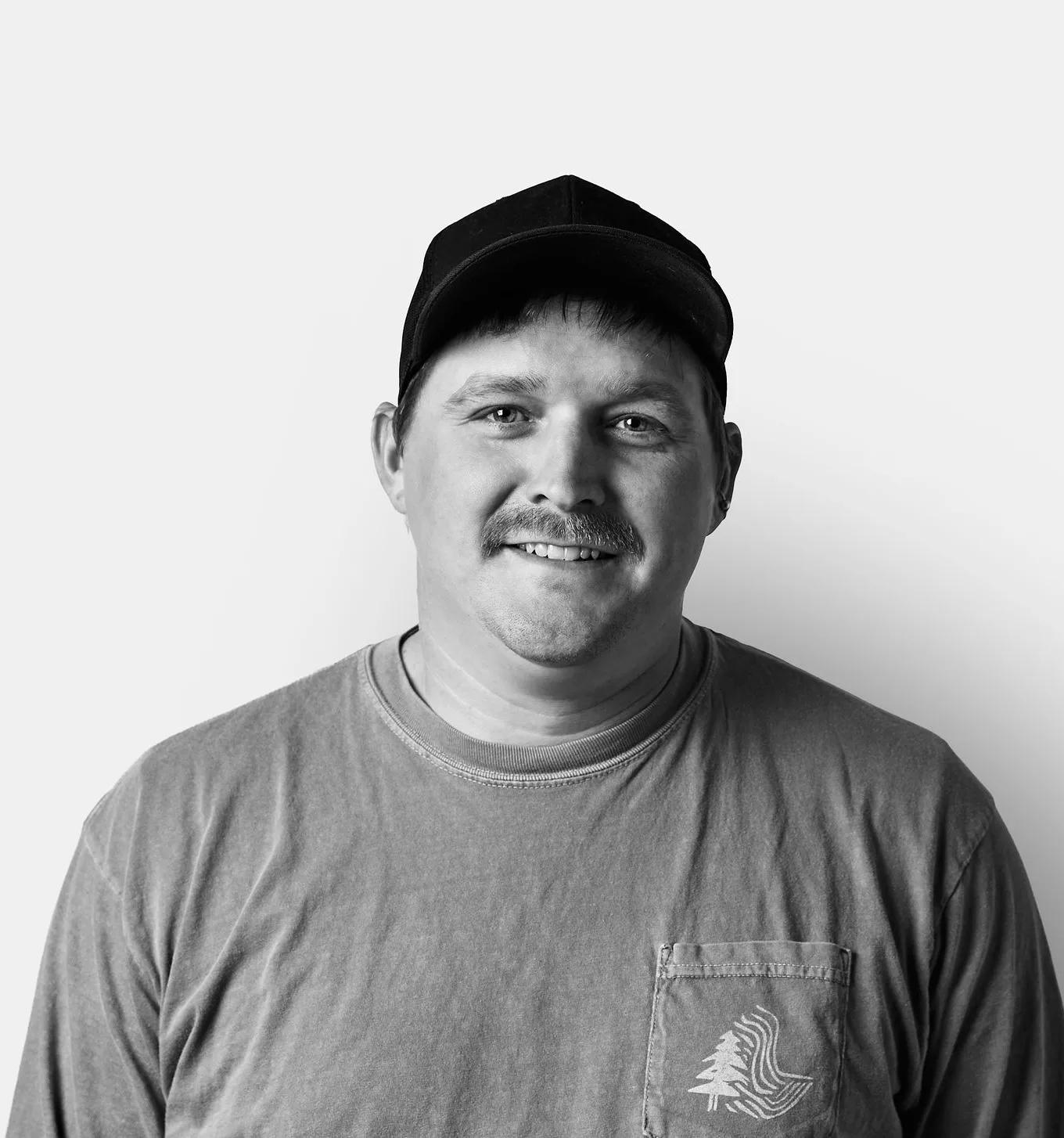 A man in a long-sleeve shirt and cap smiles at the camera against a plain white background, shown in black and white.
