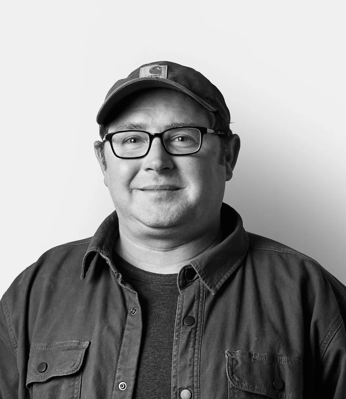 A man wearing glasses, a cap, and a button-up jacket stands smiling against a plain white background.