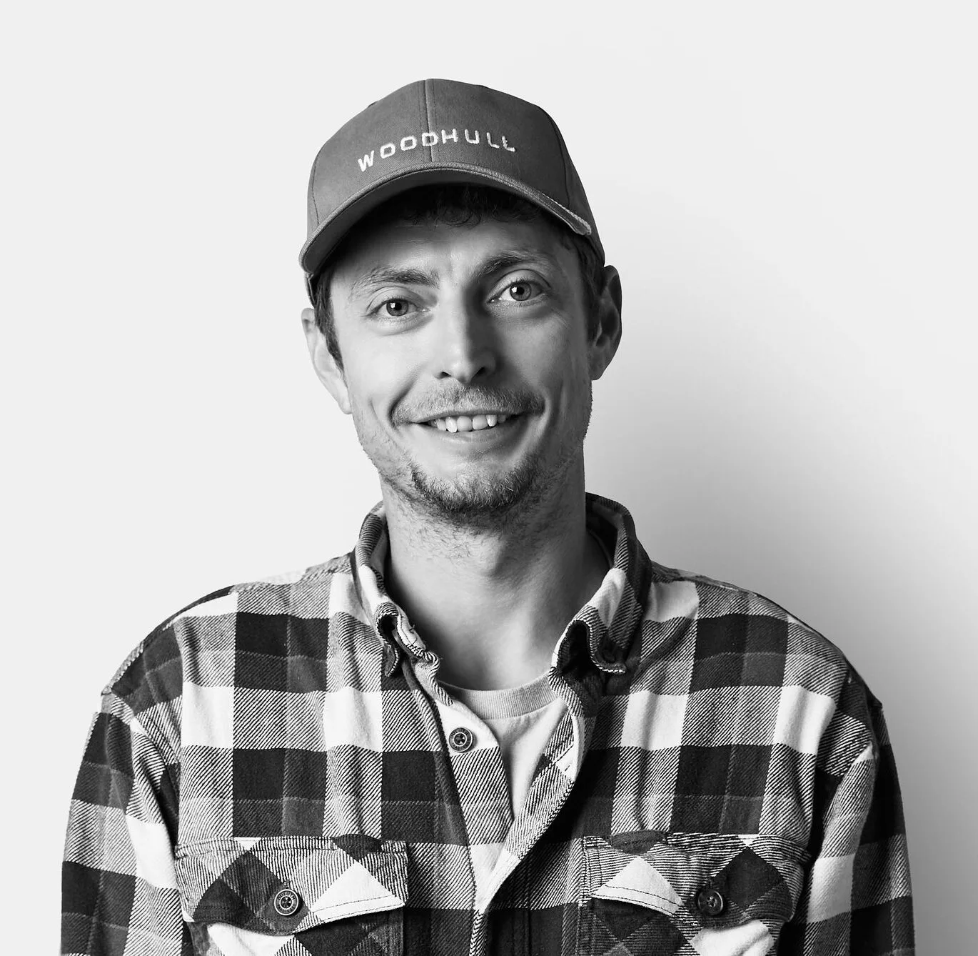 A man wearing a plaid shirt and a cap labeled WOODHULL smiles at the camera against a plain, light background.