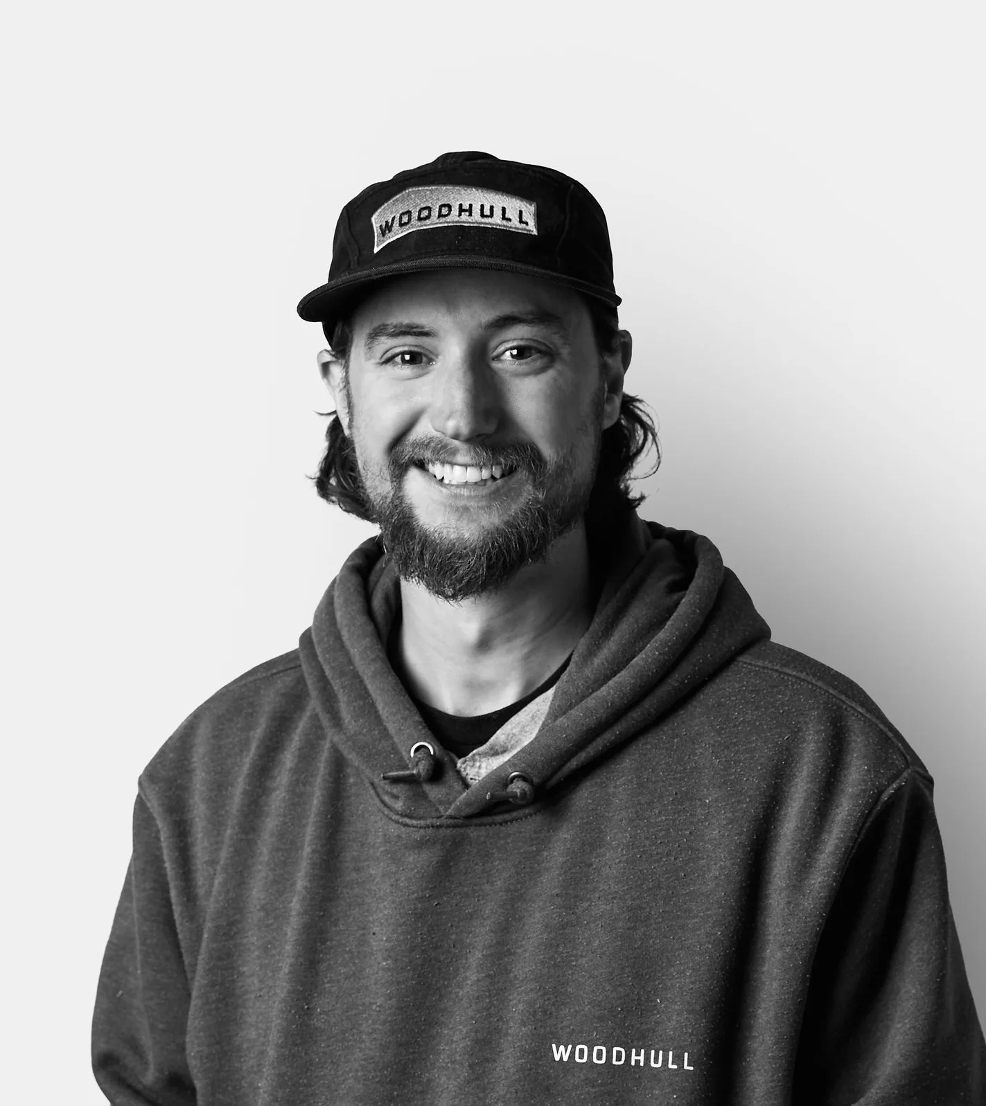 A man with a beard and mustache smiles, wearing a WOODHULL cap and hoodie, standing against a plain white background.