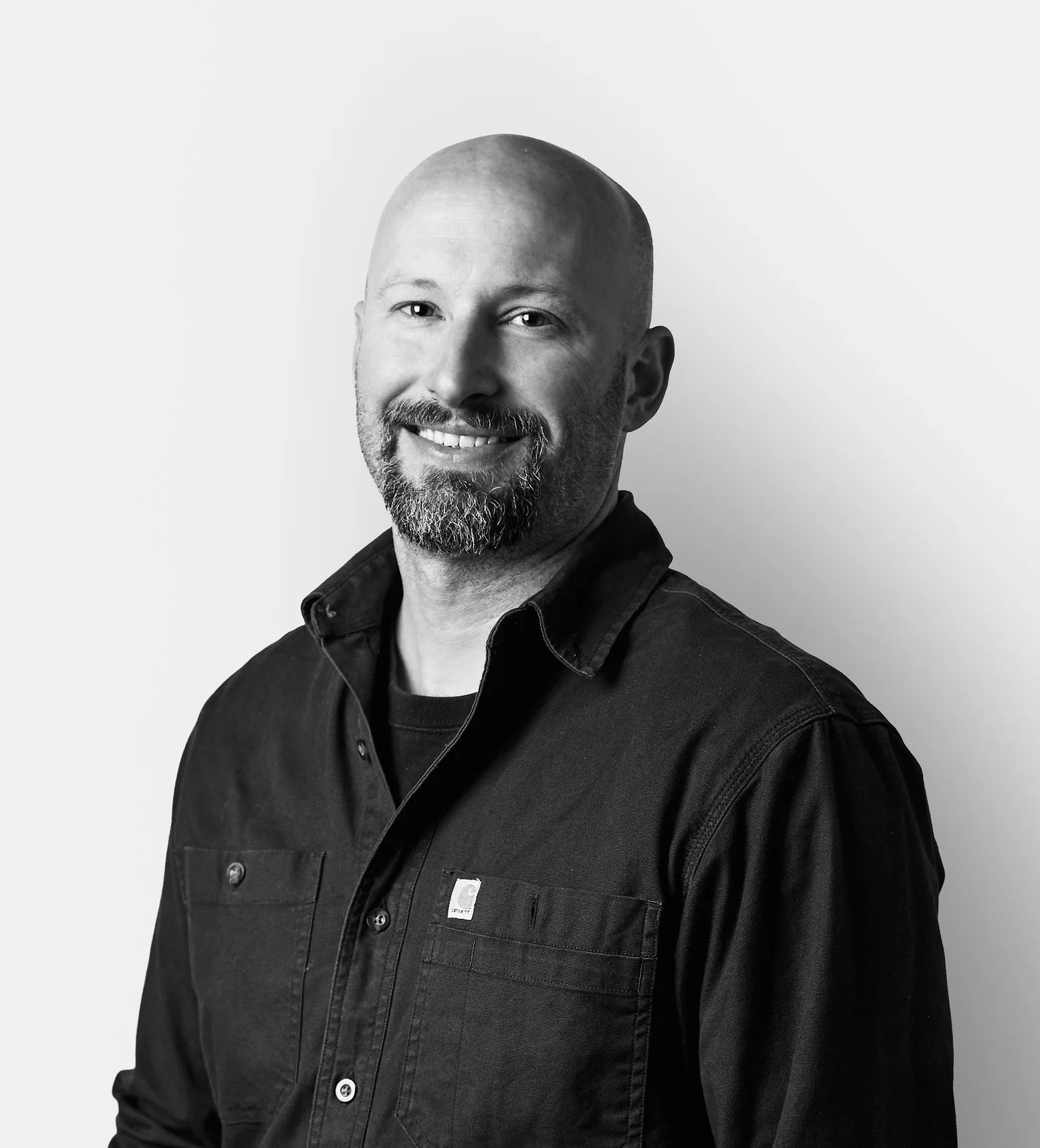 A smiling man with a bald head and beard wearing a dark button-up shirt stands against a plain, light background.