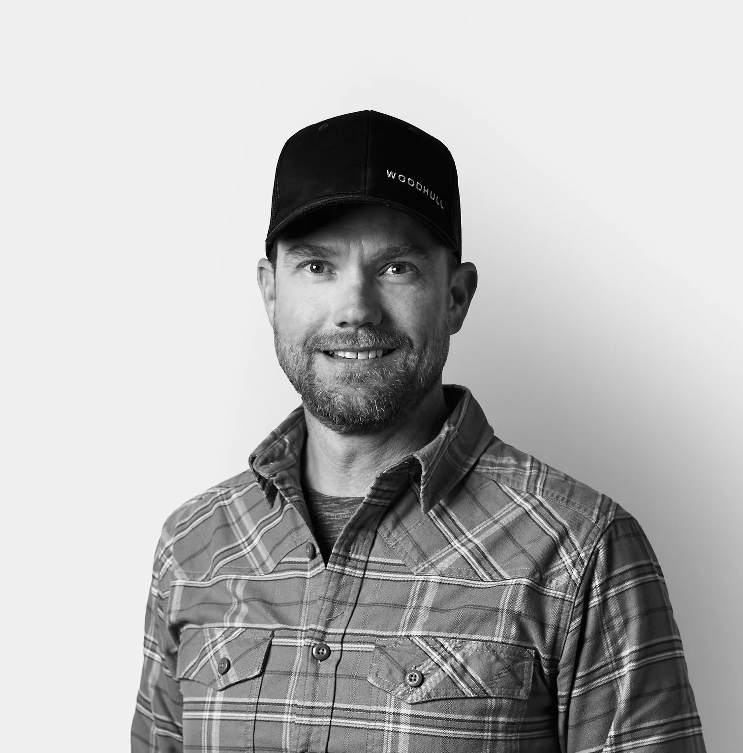 Man in a plaid shirt and a black cap smiling, standing against a plain white background.