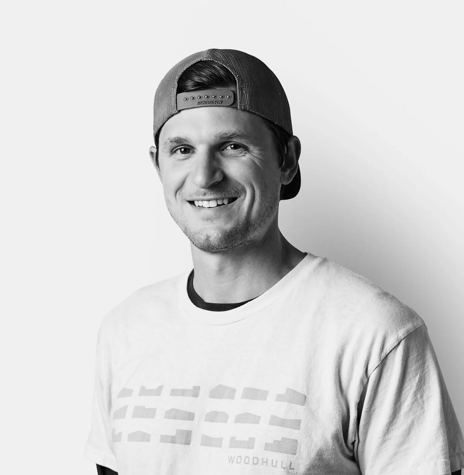 Smiling young man wearing a backwards baseball cap and a T-shirt over a long-sleeve shirt, standing against a plain white background.