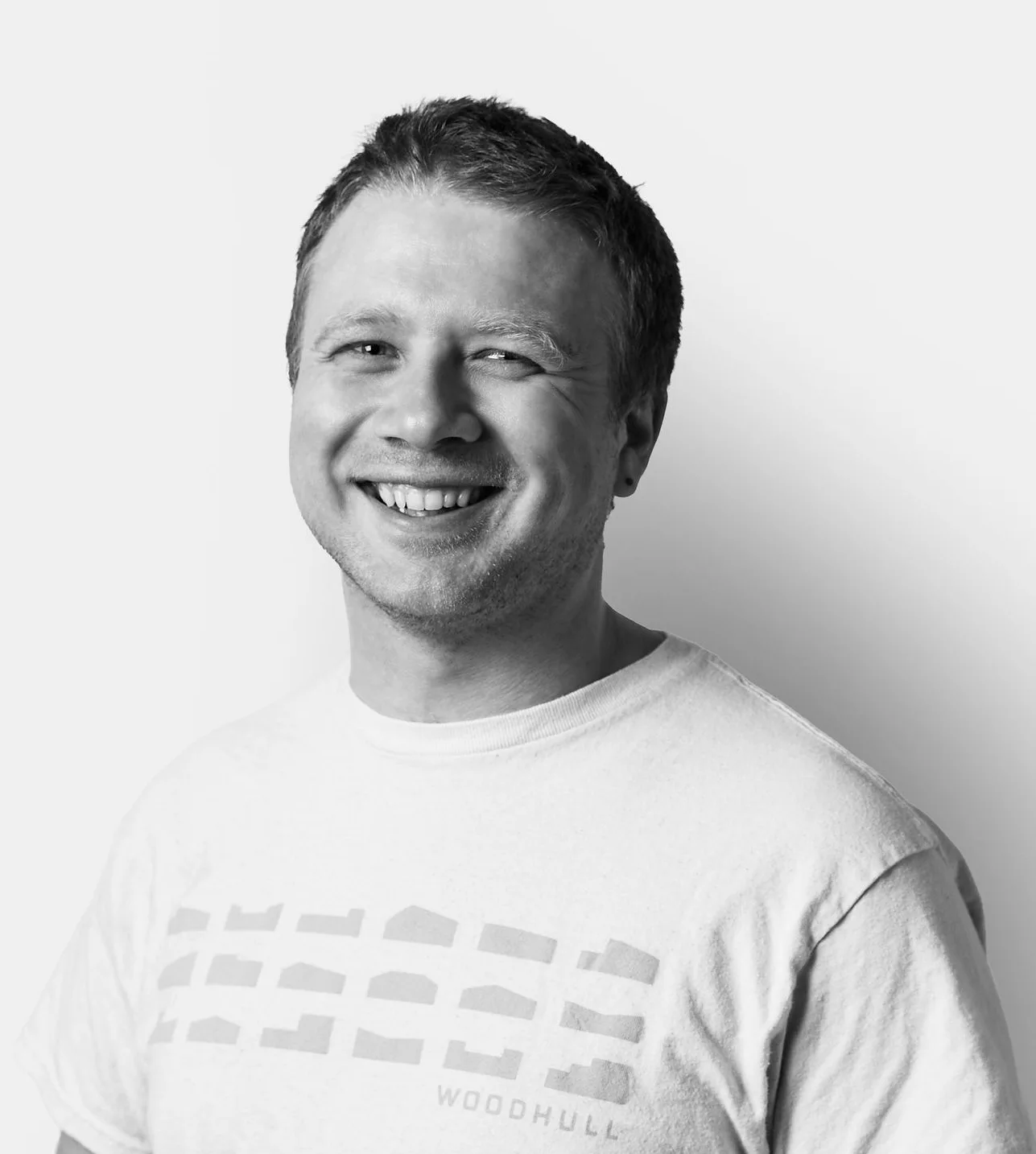 Smiling man in a light-colored Woodhull t-shirt stands against a plain white background, looking directly at the camera.