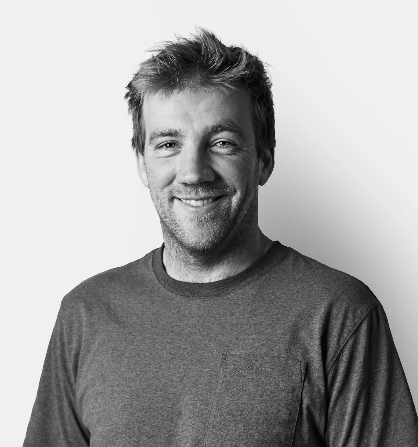 A smiling man with tousled hair wears a grey t-shirt and stands against a plain white background.