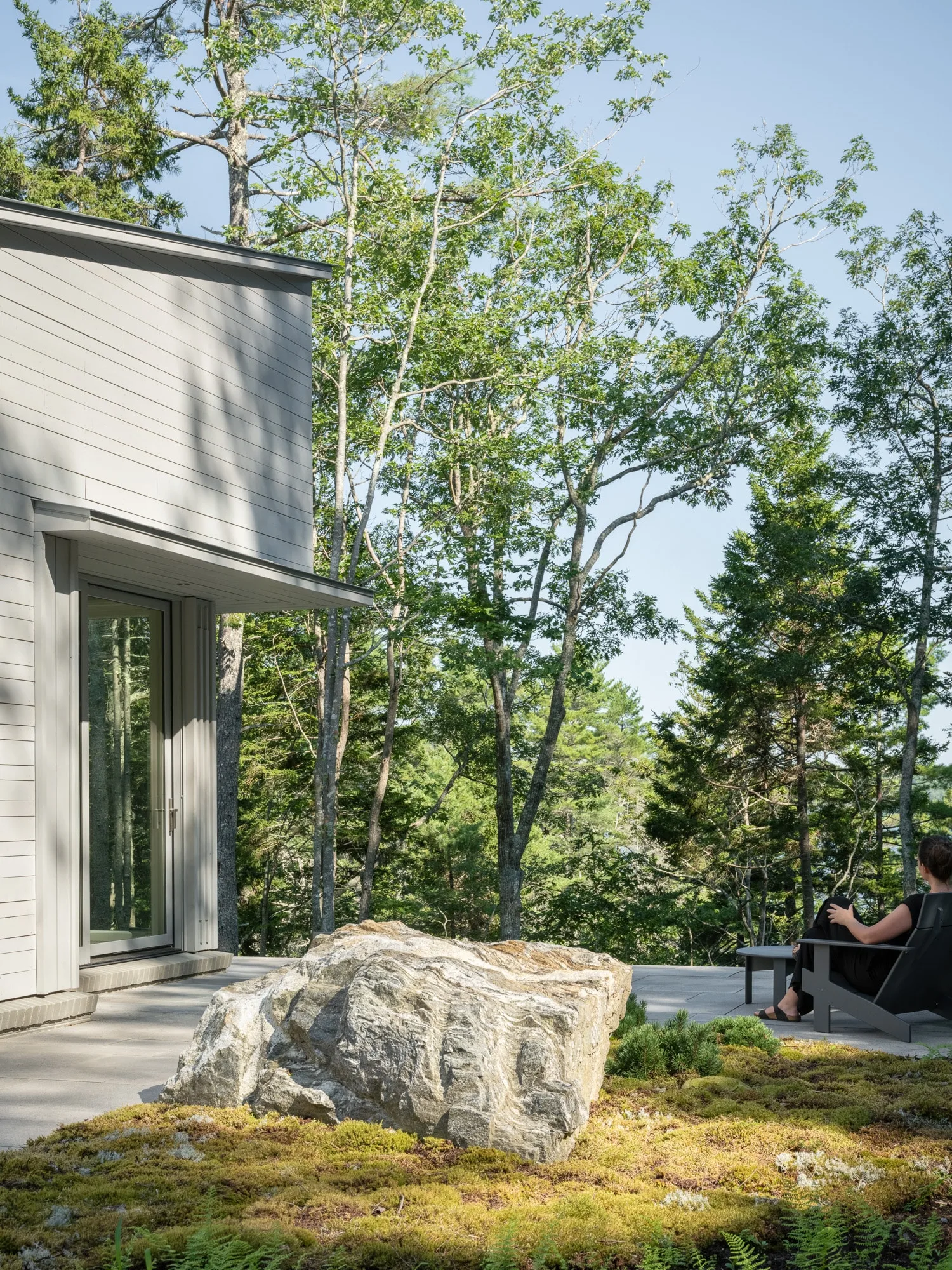 Outdoor patio of a luxury Maine home, surrounded by moss-covered rocks, trees, and modern Adirondack chairs for enjoying the tranquil forest setting